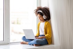 Cute black little girl with wireless headphones using laptop at home, sitting on window and looking at screen, typing on computer keyboard, playing video games on computer, copy space