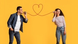 Message With Love. Happy Young Arab Man Telling Romantic Words To His Girlfriend Through Tin Phone With Drawn Red Heart As String, Loving Middle Eastern Couple Posing Over Yellow Background