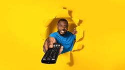 Cheerful african american man watching TV and pointing television remote controller to camera, switching channels, posing in torn yellow paper hole. Happy black male enjoying movie