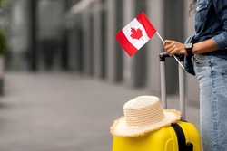 Cropped of woman traveller holding yellow luggage with straw hat on and flag of Canada, going vacation overseas, airport background, copy space. Immigration, education abroad concept