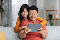 Happy asian lovers spending time together at home, positive middle aged man and young woman relaxing on couch in living room, hugging and using digital tablet, playing mobile game, copy space