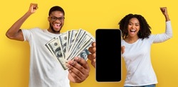 Giveaway Concept. Portrait of overjoyed young black couple holding a lot of dollar cash and showing smartphone with blank screen, posing with bunch of money over yellow studio background, mockup