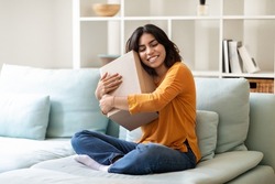 Joyful Young Arab Woman Embracing Big Cardboard Box While Sitting On Couch At Home, Happy Middle Eastern Female Holding Delivery Parcel And Smiling, Enjoying Making Online Purchases, Copy Space