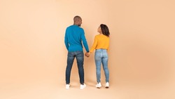 Full length of black man and woman standing with their backs to camera, looking at each other and holding hands against peach studio wall. African american couple expressing affection