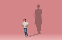 Childhood dreams concept. Cool cheerful school aged asian girl dreaming to become a doctor, shadow of female doc behind her, kid posing over pink wall studio background, collage, copy space
