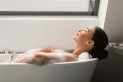 Relaxed Female Taking Bath With Foam Lying In Bathtub In Modern Bathroom Indoors. Woman Bathing Enjoying Bodycare Routine With Eyes Closed At Home. Spa And Wellness. Side View, Selective Focus