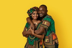 Portrait of happily married black couple in bright national african costumes embracing and smiling at camera, standing against yellow studio background, copy space. Ethnicity, culture concept