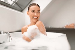 Smiling Lady Taking Relaxing Bath Looking Aside Sitting In Bathtub With Foam In Modern Bathroom At Home. Woman Bathing Enjoying Bodycare Routine. Beauty And Spa Concept