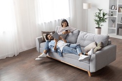 High angle view of relaxed cheerful stylish millennial eastern lovers man and woman chilling on couch in cozy living room, using gadgets at home, scrolling on social media, reading online, copy space