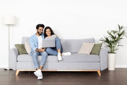 Loving happy beautiful eastern couple in stylish casual outfit sitting on couch at home, embracing, using laptop, young indian man and woman watching movie online, copy space, full length