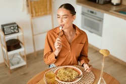 Young lady eating delicious homemade pasta, enjoying tasty lunch with closed eyes while sitting at table in light cozy kitchen interior, free space, above view