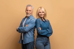 Partnership, teamwork concept. Cheerful attractive confident senior man and woman wearing casual denim outfit standing back to back with arms crossed over beige studio background, smiling at camera