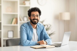Successful Freelancer. Happy Millennial Indian Man Sitting At Desk With Laptop, Handsome Eastern Guy Smiling And Posing At Camera While Working On Computer At Home Office, Copy Space