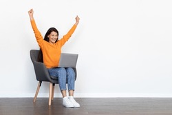 Thrilled emotional beautiful millennial hindu woman wearing casual outfit sitting in armchair over white blank wall, looking at computer laptop screen, gesturing raising hands up, copy space
