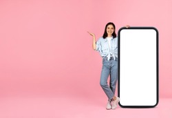 Happy cheerful pretty young woman wearing casual outfit posing with huge smartphone with white blank screen and gesturing, copy space, mockup on pink background. Nice online deal, offer
