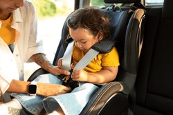 Asian Mom Sitting Down Her Toddler Daughter In Baby Car Seat, Adjusting Harness Straps For Safety During Automobile Journey. Cropped Shot, Closeup Of Child In Chair For Safe Auto Trip