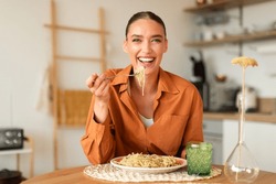 Happy young european lady eating delicious homemade Italian pasta, enjoying tasty lunch, looking at camera and laughing, sitting in cozy kitchen interior, copy space