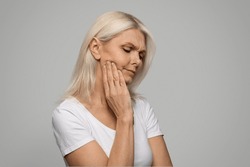 Closeup of sad mature woman touching her cheek, rubbing aching jawline, upset senior lady having tooth pain, suffering strong toothache, standing over grey studio background, copy space