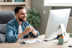 Happy Young Businessman Drinking Coffee At Workplace And Using Computer, Handsome Male Entrepreneur Sitting At Work Desk, Looking At Monitor Screen And Smiling, Enjoying Casual Day In Office