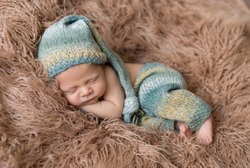 Little baby weared in light blue-yellow knitted beanie and pants sweetly sleeping on the light brown soft coverlet