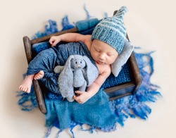 Little smiling baby weared in a blue knitted beanie sweetly sleeping in the small brown bed covered with blue-gray blankets