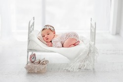 Beautiful newborn baby girl wearing lace costume ana wreath lying on her tummy on stylized white bed during studio photoshoot. Cute portrait of infant child in white room