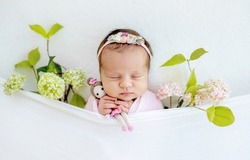 Newborn baby girl holding knitted doll and sleeping wearing wreath. Adorable infant child kid napping with flower plant decoration