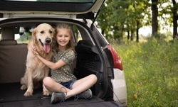 Preteen girl sitting with golden retriever dog in the car trunk and smiling looking at the camera. Child kid hugging purebred doggy pet in the vehicle at the nature