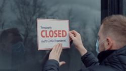 Caucasian male wearing medical mask puts a Temporary closed due COVID-19 pandemic sign on a window. Coronavirus pandemic, small business shutdown