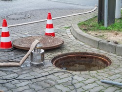 Round manhole cover next to a water meter shaft in Berlin with a pump, water hose and shut-off pylons in front of it.