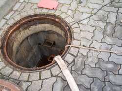Opened round water meter shaft with a water hose leading in and a tether to drain the water after a pipe break on a public walkway in Berlin.