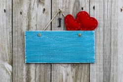 Country red fabric hearts and black iron house key hanging on antique teal blue blank rustic wooden sign; Valentine's Day background with painted wood copy space