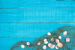 Collection of white seashells in fish net border on antique rustic teal blue wood background; blank beach sign with painted copy space