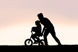 A Silhouette of a good father is helping his young girl child learn to ride her bicycle with training wheels on a summer day.