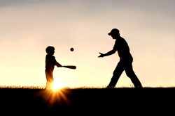 A silhouette of a father and his young child playing baseball outside, isolated against the sunsetting sky on a summer day.