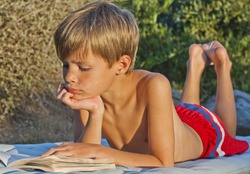 Cute, young boy reading a book in the sunshine on a sun lounger