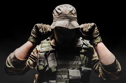 Photo of a fully equipped camouflaged soldier putting on panama hat with tactical vest and gloves  on dark background wirh fog.
