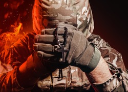 Photo of a military soldier in uniform and armor helmet sitting and holding cross religious necklace on back background with red fire burning glow.