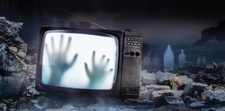 Horror photo of an old black  scary haunted tv set with ghost hands on screen, standing on dark foggy ruined city with spirit figures background.