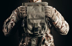 Photo of soldier in camouflaged uniform and tactical gloves holding leg bag on black background close-up view.