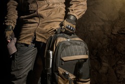Backpack and gear composition.Man in storm jacket and tactical military gloves holding a backpack with travel gear on stone wall background.