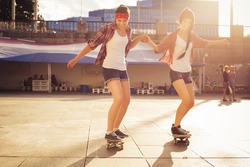 Two Brunette teenage girls friends in hipster outfit (jeans shorts, gumshoes, plaid shirt, hat) with a skateboard at the park outdoors. Copy space