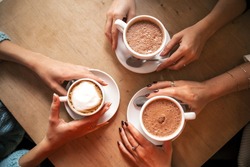 Flat lay image from above: three hands holding three cup of coffee on a wooden table. Cozy, lifestyle image