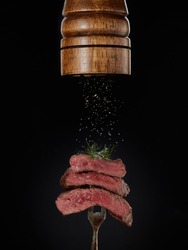 Roasted meat. Milled spices falling from pepper mill on grilled pieces of beef steak medium rare on fork on black background. Steak menu. Grilled menu.