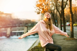cool hipster young girl in pink hoodie standing by a river in the city enjoying the sun of autum or springtime
