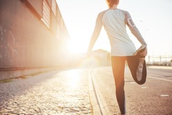 Athlete woman preparing for running on the city street. Legs warming and stretching. Sport tight clothes. Bright sun, blurry background. Horizontal
