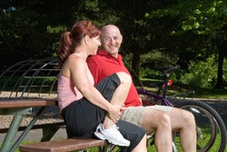 Happy couple sitting on a park bench next to their bicycles. They are looking at each other and smiling. Horizontally framed photograph