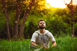 A bearded man is meditating on green grass in the park with face raised up to sky and eyes closed on sunny summer day. Concept of meditation, dreaming, wellbeing and healthy lifestyle