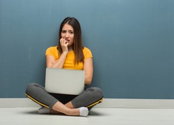 Young woman sitting on the floor with a laptop biting nails, nervous and very anxious