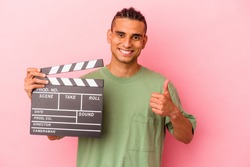 Young venezuelan man holding a clapperboard isolated on pink background smiling and raising thumb up